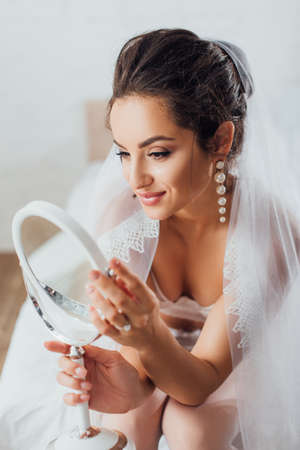 Selective focus of bride in bra and veil looking at mirror in bedroomの写真素材