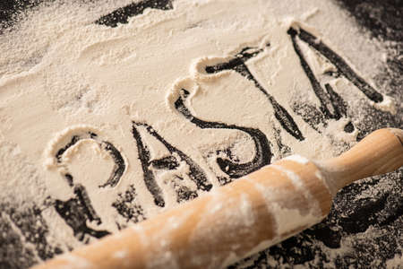 pasta lettering written on flour near rolling pin on black background
