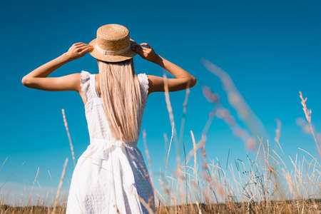 back view of woman in white dress touching straw hat while standing in field against blue sky,...