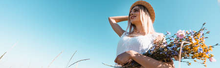 low angle view of young woman touching straw hat while holding wildflowers and looking away, panoramic shotの写真素材