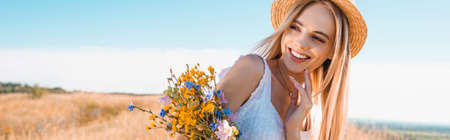 panoramic shot of blonde woman in straw hat holding wildflowers and looking away against blue sky