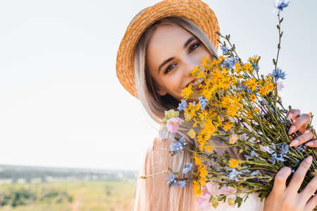 blonde woman in straw hat holding wildflowers while looking at camera against clear skyの写真素材