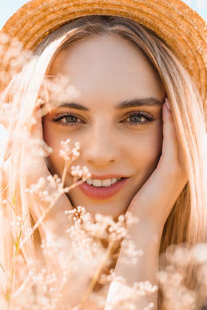 portrait of sensual blonde woman in straw hat looking at camera and touching face near wildflowers
