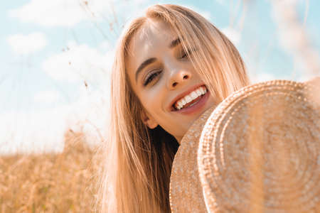 selective focus of excited blonde woman holding straw hat and looking at camera near grass in meadowの写真素材