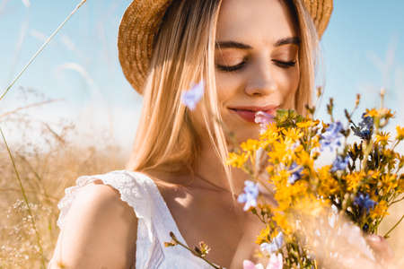selective focus of sensual woman in straw hat smelling bouquet of wildflowers with closed eyesの写真素材