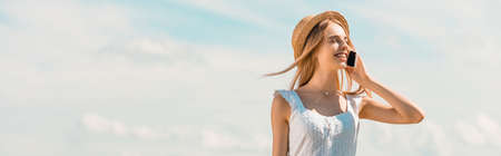 panoramic shot of blonde woman in straw hat talking on smartphone with closed eyes against blue sky