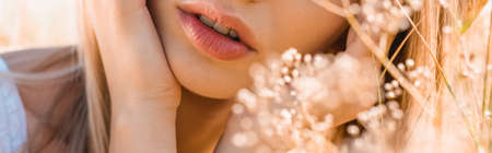 partial view of young woman touching face near wildflowers, panoramic shot