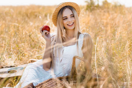 selective focus of woman in white dress and straw hat holding ripe apple while sitting in fieldの写真素材