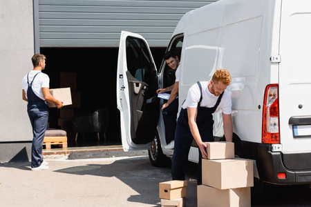 Selective focus of loader in overalls taking cardboard box while colleagues working near truck on urban streetの写真素材
