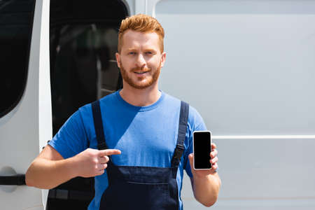Loader in overalls pointing at smartphone with blank screen near truck outdoors