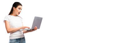 panoramic crop of shocked brunette woman in white t-shirt holding laptop isolated on whiteの写真素材