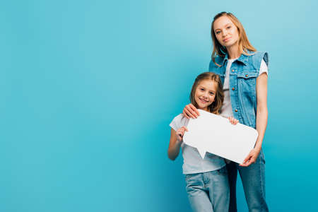young mother in denim clothes holding speech bubble with daughter isolated on blue