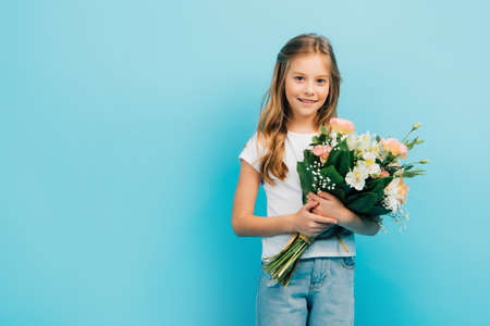 child in white t-shirt and blue jeans looking at camera while holding bouquet isolated on blue
