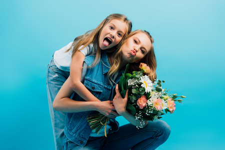 girl sticking out tongue and hugging mother pouting lips while holding bouquet of flowers isolated on blueの写真素材