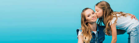 horizontal image of young woman looking at camera while daughter kissing her isolated on blueの写真素材
