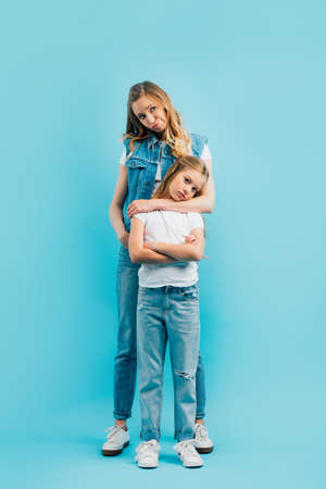 full length view of upset woman in denim clothes hugging offended daughter in white t-shirt and jeans on blueの写真素材