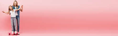 horizontal image of mother supporting daughter standing on penny board by holding her hands on pink