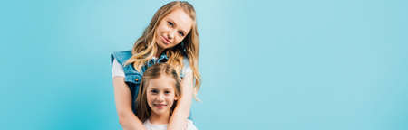 horizontal image of young woman hugging daughter while looking at camera isolated on blue