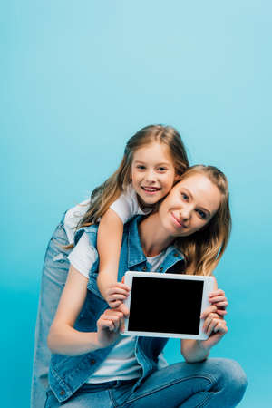 mother and daughter in denim clothes showing digital tablet with blank screen and looking at camera isolated on blueの写真素材