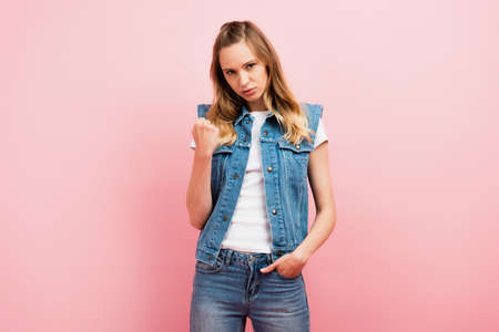 irritated woman in denim clothes showing clenched fist while standing with hand in pocket isolated...