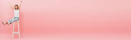 girl in white t-shirt and blue jeans sitting on high chair with outstretched legs and showing winner gesture on pink, panoramic shotの写真素材