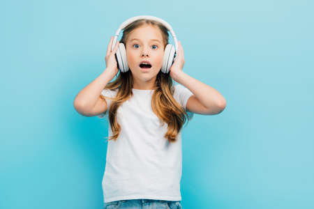 excited girl in white t-shirt touching wireless headphones while looking at camera isolated on blueの写真素材
