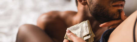 Panoramic crop of woman holding dollars and touching chin of shirtless man on bedの写真素材