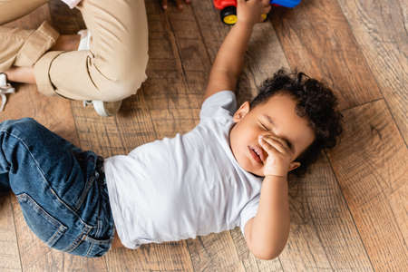 top view of sleepy african american boy touching eyes while lying on floor at homeの写真素材