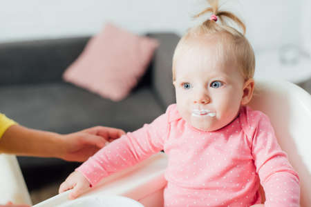 Selective focus of baby girl with messy mouth sitting on feeding chair near mother at homeの写真素材