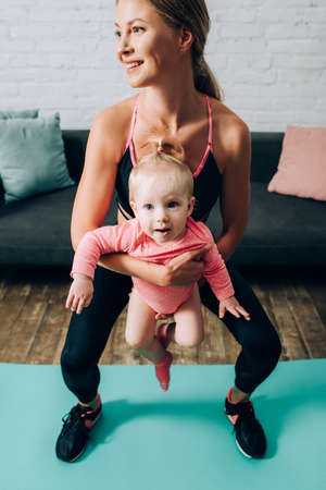 Mother in sportswear holding baby girl while training on fitness mat at homeの写真素材