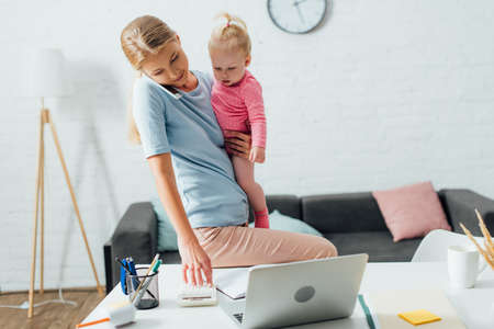 Selective focus of woman talking on smartphone and using calculator while holding daughter at home