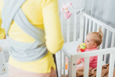 Selective focus of infant girl biting toy in crib near mother with sling at home