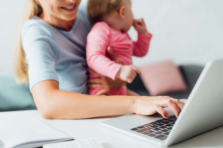 Selective focus of freelancer using laptop while holding baby daughter at homeの写真素材