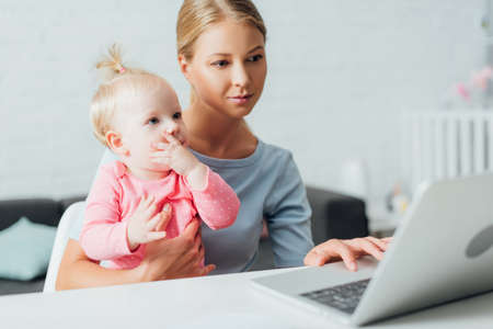 Selective focus of woman holding daughter and using laptop at table
