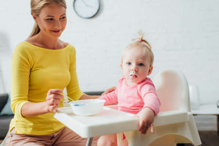 Selective focus of baby girl looking at camera near mother with spoon and bowl of baby food at homeの写真素材