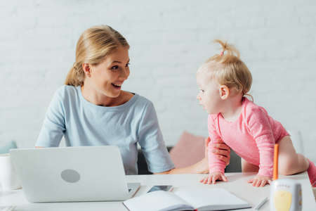 Selective focus of woman looking at daughter while working with laptop near baby monitor on tableの写真素材