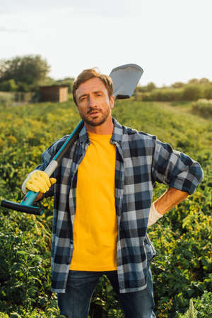 farmer in plaid shirt looking at camera while standing in field with shovel