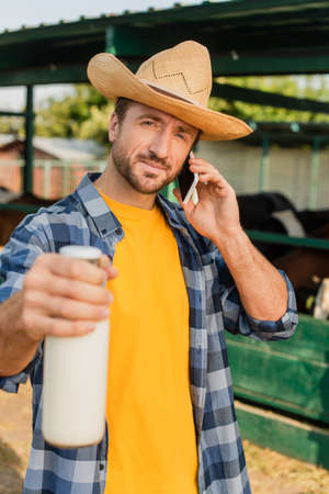 selective focus of rancher in straw hat and checkered shirt talking on smartphone while showing bottle of milkの写真素材
