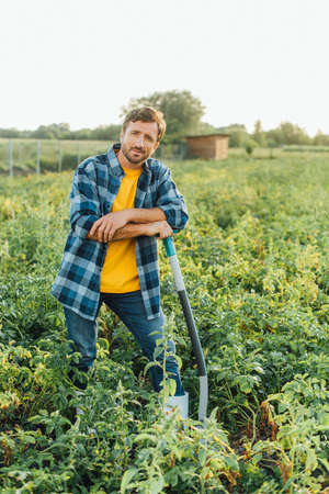 farmer in checkered shirt looking at camera while leaning on shovel in field