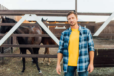 rancher in plaid shirt standing near corral with horses and looking at cameraの写真素材