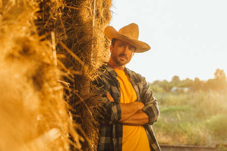 selective focus of farmer in plaid shirt leaning on hay stack with crossed arms while looking at cameraの写真素材