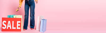 Panoramic crop of woman holding credit card near card with sale lettering and purchases on pink backgroundの写真素材