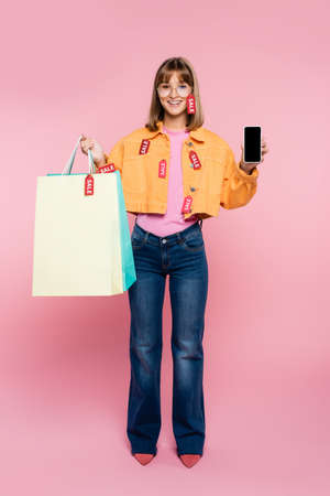 Young woman with price tags on jacket holding shopping bags and smartphone with blank screen on pink...