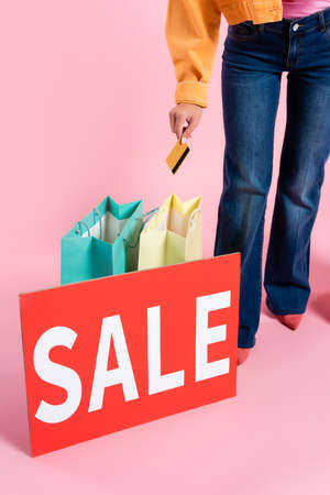 Cropped view of woman holding credit card near shopping bags and card with sale lettering on pink...