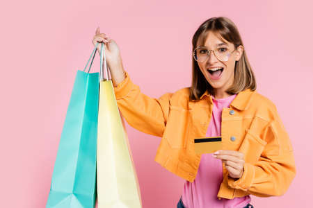 Excited woman in yellow jacket and sunglasses holding colorful shopping bags and credit card on pink...