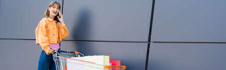 Panoramic shot of young woman talking on smartphone near cart with shopping bags and buildingの写真素材