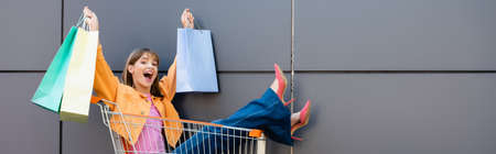 Horizontal image of excited woman holding colorful shopping bags while sitting in cart near buildingの写真素材