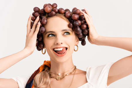 rustic blonde woman holding grapes on head and showing tongue isolated on whiteの写真素材
