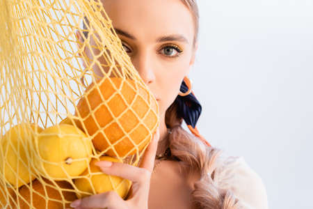 rustic blonde woman posing with citrus fruits in yellow string bag in front of face isolated on...