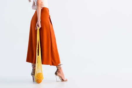 cropped view of woman in skirt posing with citrus fruits in yellow string bag isolated on whiteの写真素材